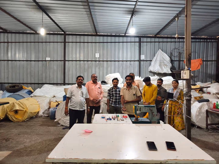 Team standing around a stitching machine in the plant, with rolls of woven fabric stacked behind them