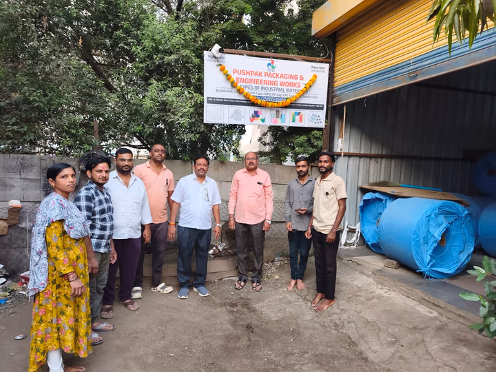 Company signboard with a marigold garland above a group of people, with blue rolls of woven fabric beside them